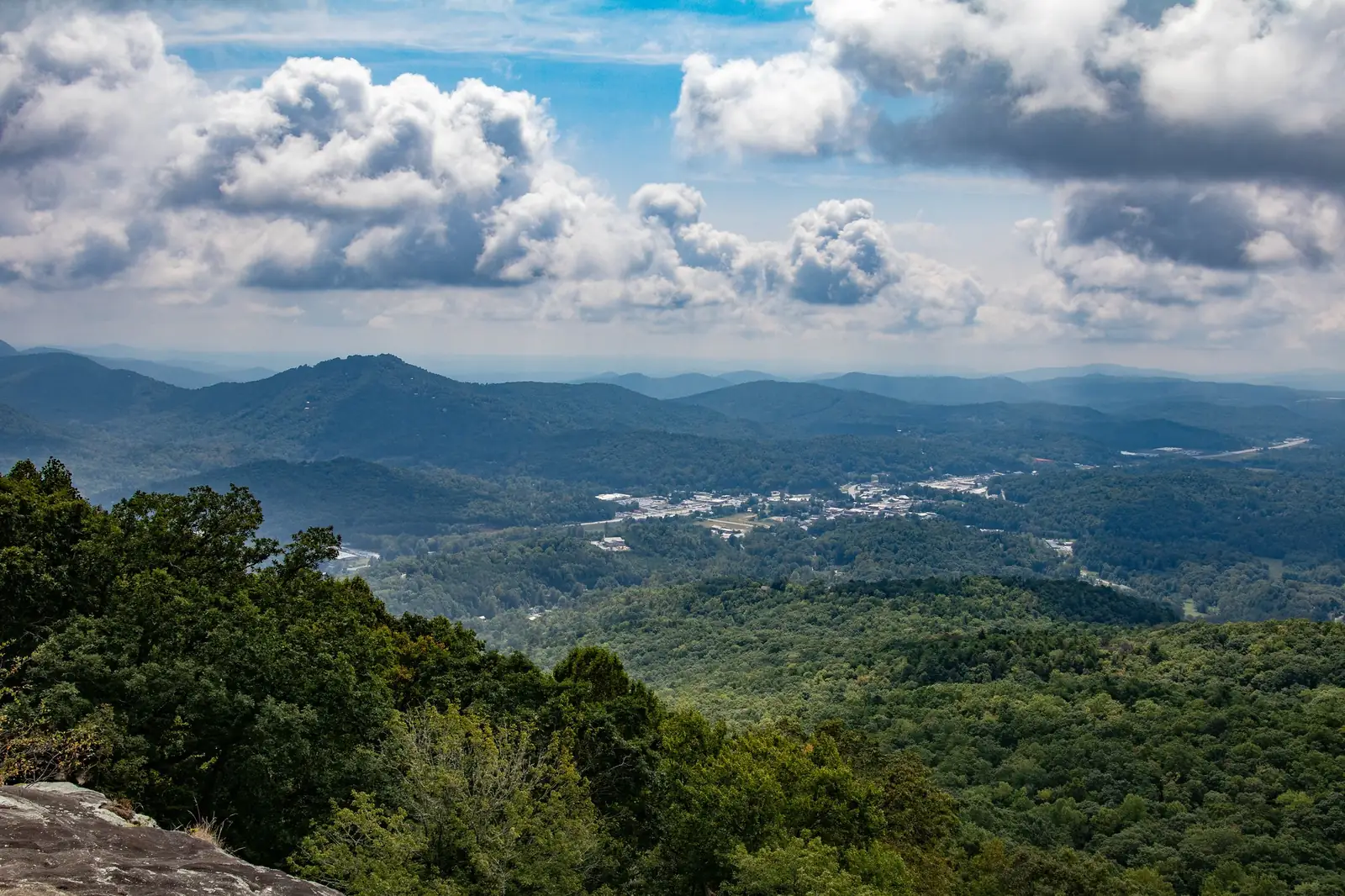 Wide mountain panorama from a rocky overlook: forested slopes in the foreground, a town in the valley below, a peaked mountain at left, and towering white clouds in a blue sky.