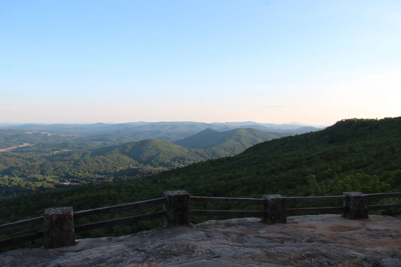 A stone-post and timber railing on a bare granite overlook in the foreground, looking out over a forested slope, rolling green hills and a distant valley under a clear pale-blue sky.