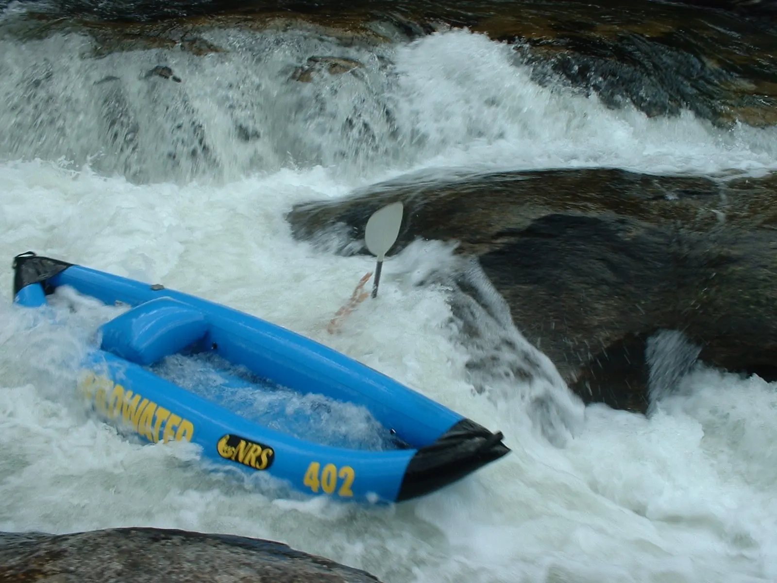 Close view of churning white water pouring over dark rocks, with an empty blue inflatable kayak marked "402" tipped sideways in the rapid and a paddle blade sticking up.
