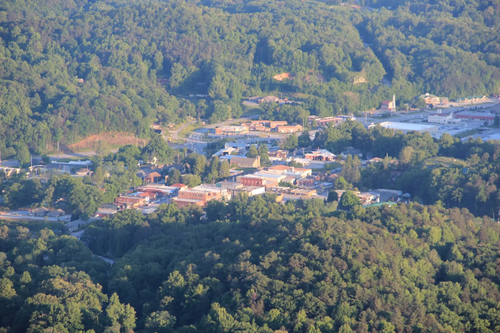 Telephoto view from a mountaintop down onto a small town nestled in a forested valley, with brick commercial buildings, a white steeple and wooded hills on every side in early-autumn color.