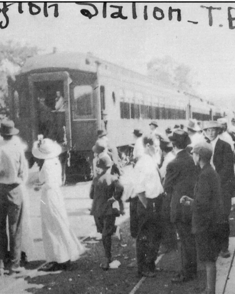 Black-and-white historic photograph of a crowd of men in hats and women in long white dresses gathered on a station platform beside a passenger railcar; "Clayton Station - T.F. Ry" is handwritten across the top.
