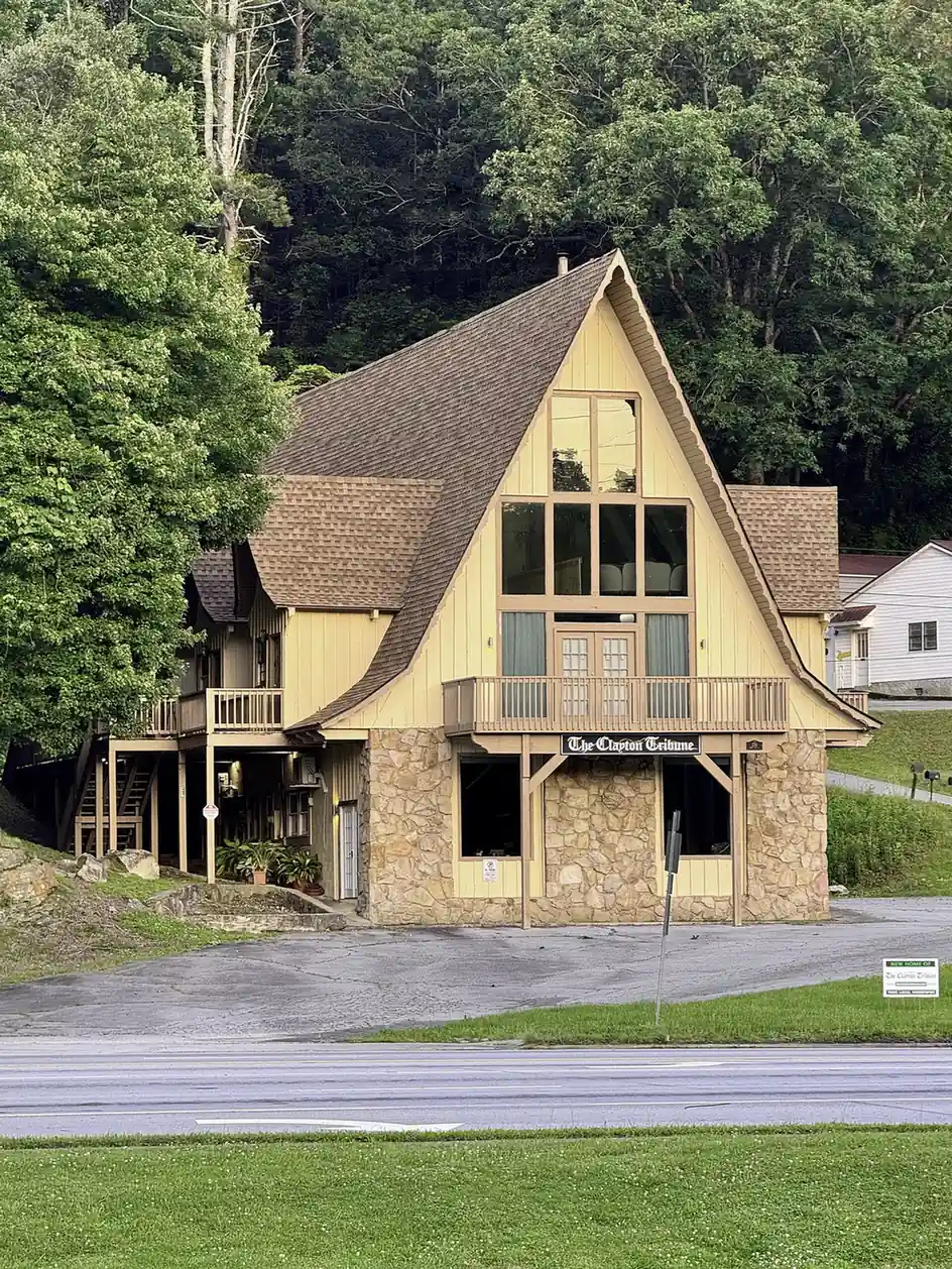 A steep A-frame building with cream board-and-batten siding, a stone lower story, a wooden balcony and a black sign reading "The Clayton Tribune", set against a wooded hillside above an empty paved lot; a small yard sign at right reads "New home of The Clayton Tribune - your local newspaper".