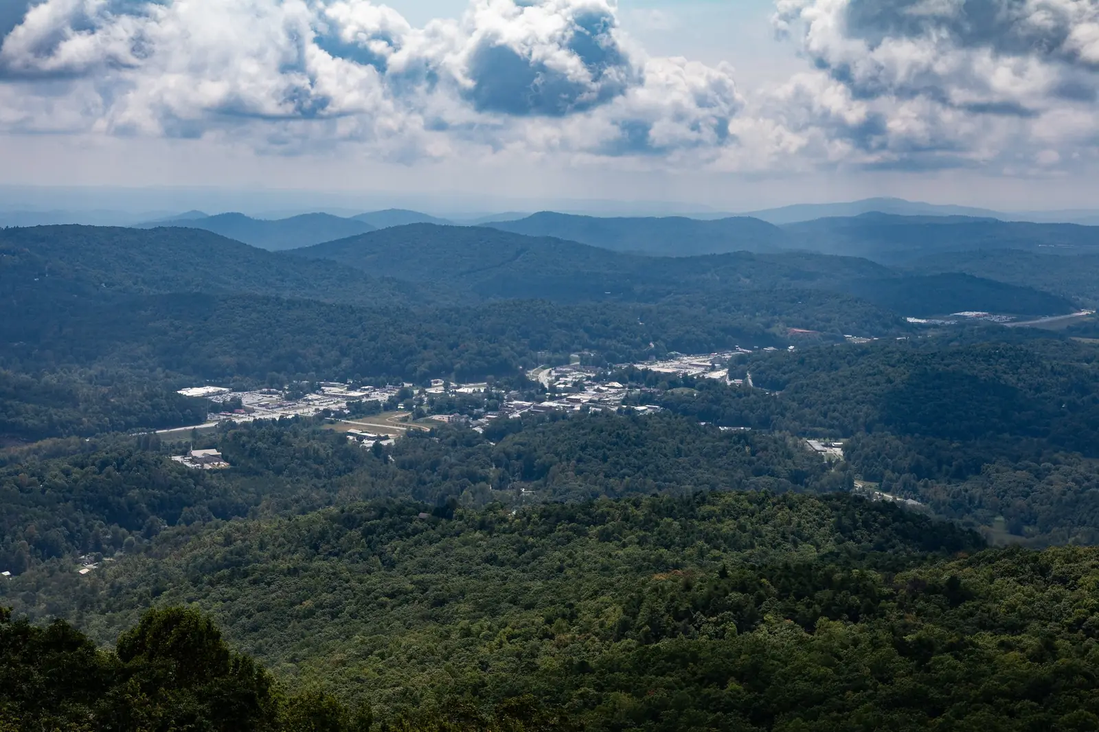View from a mountain overlook across a broad green valley with a town spread across its floor, layered blue ridges to the horizon and a band of white cumulus clouds above.