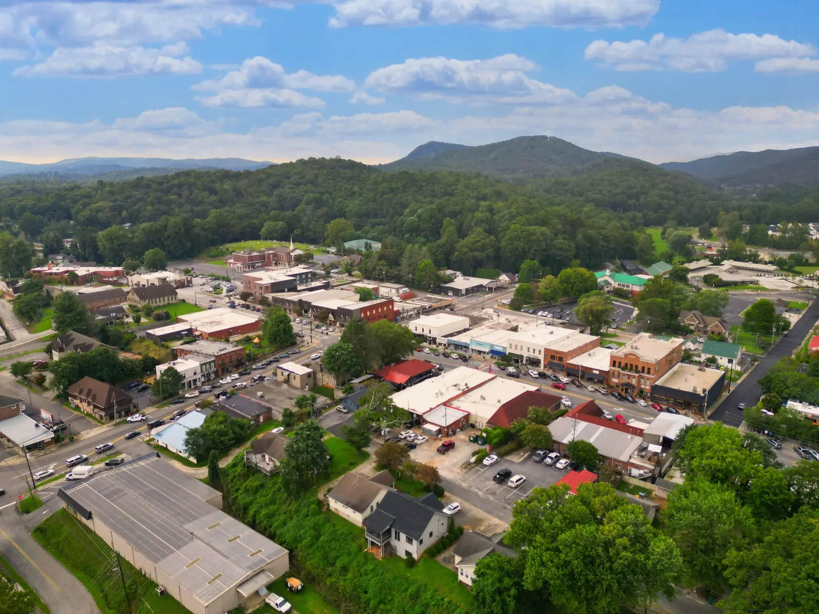 High drone view over downtown: a row of flat-roofed storefronts with colored awnings along a street lined with parked cars, an ornate brick building and red-roofed buildings in the foreground, a church steeple at left, and forested mountain ridges under a blue sky with white cumulus clouds.