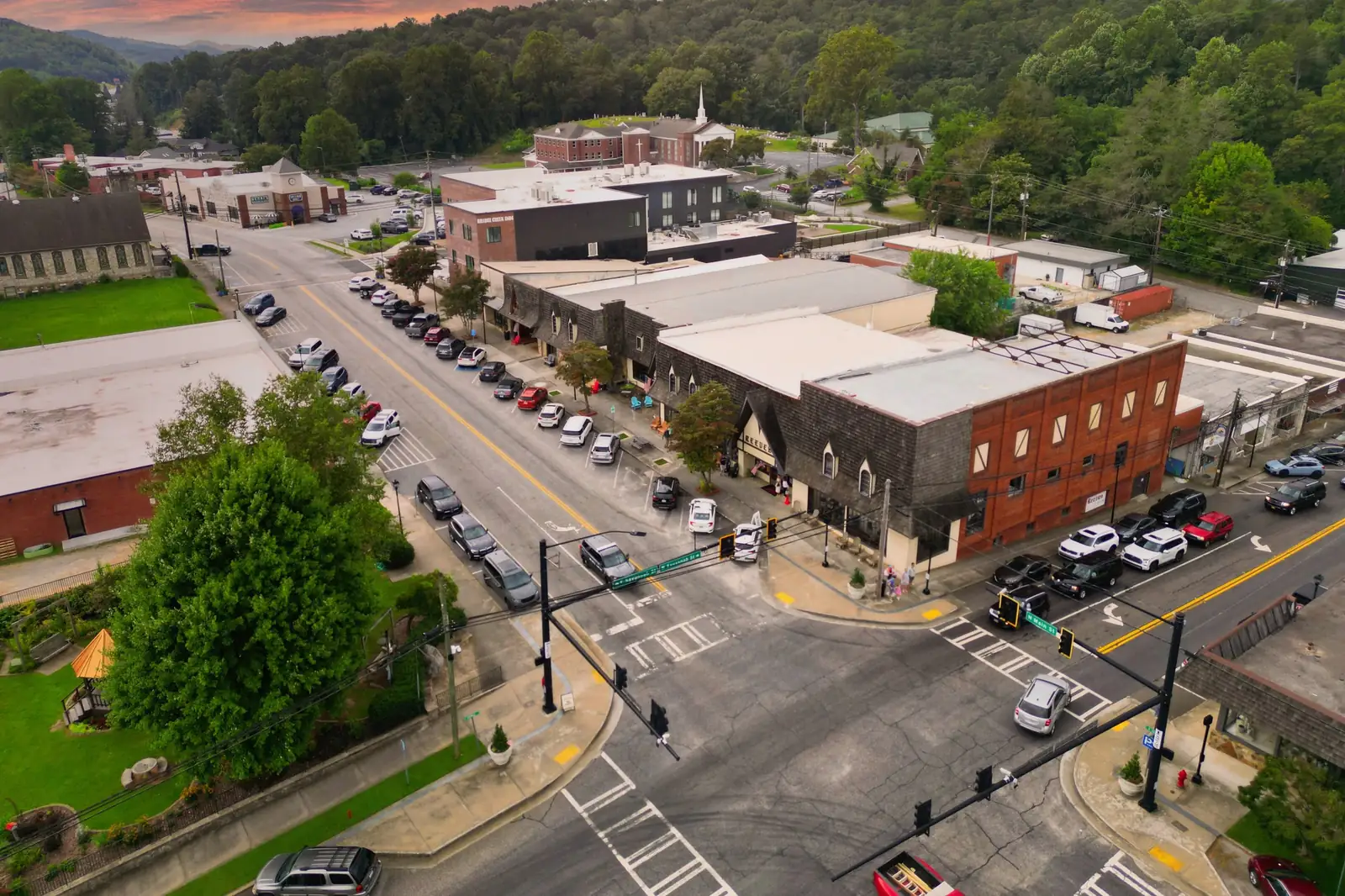 Drone view looking down on a signalized downtown crossroads with green street signs reading "N Main St" and "Savannah St"; a shingle-roofed storefront with "Reeves" lettering and a red-brick block sit on the far corner, a brick-and-black "Bridge Creek Inn" building and a white-steepled brick church with a hillside cemetery rise behind, and wooded mountains fill the top under an orange-pink evening sky.