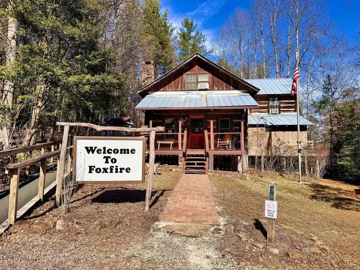 A two-story log cabin with a red door, porch and tin roof at the end of a brick walkway, a large white sign reading "Welcome To Foxfire", an American flag on a pole, and bare and evergreen trees under a deep-blue sky.