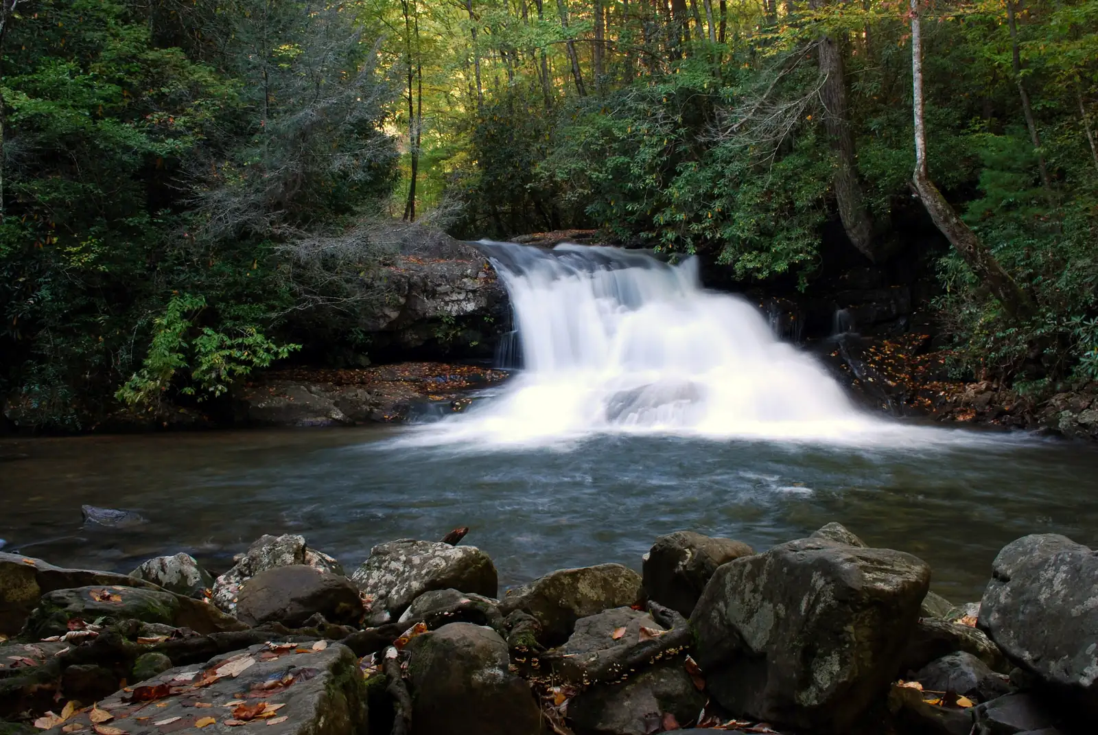 A broad white waterfall pouring over a rock ledge into a dark pool, boulders in the foreground and woods with early autumn tints around it.