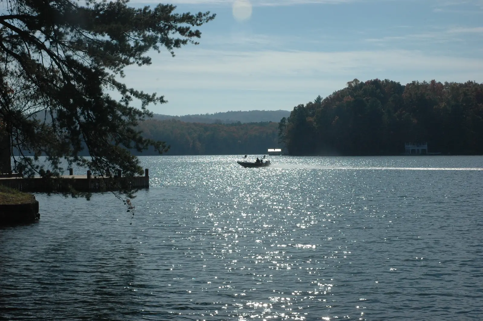 Sunlit lake water sparkling under a pale sky, framed by a dark pine bough at top left; a small fishing boat crosses mid-lake toward a wooded peninsula and distant ridges, with a dock at left.