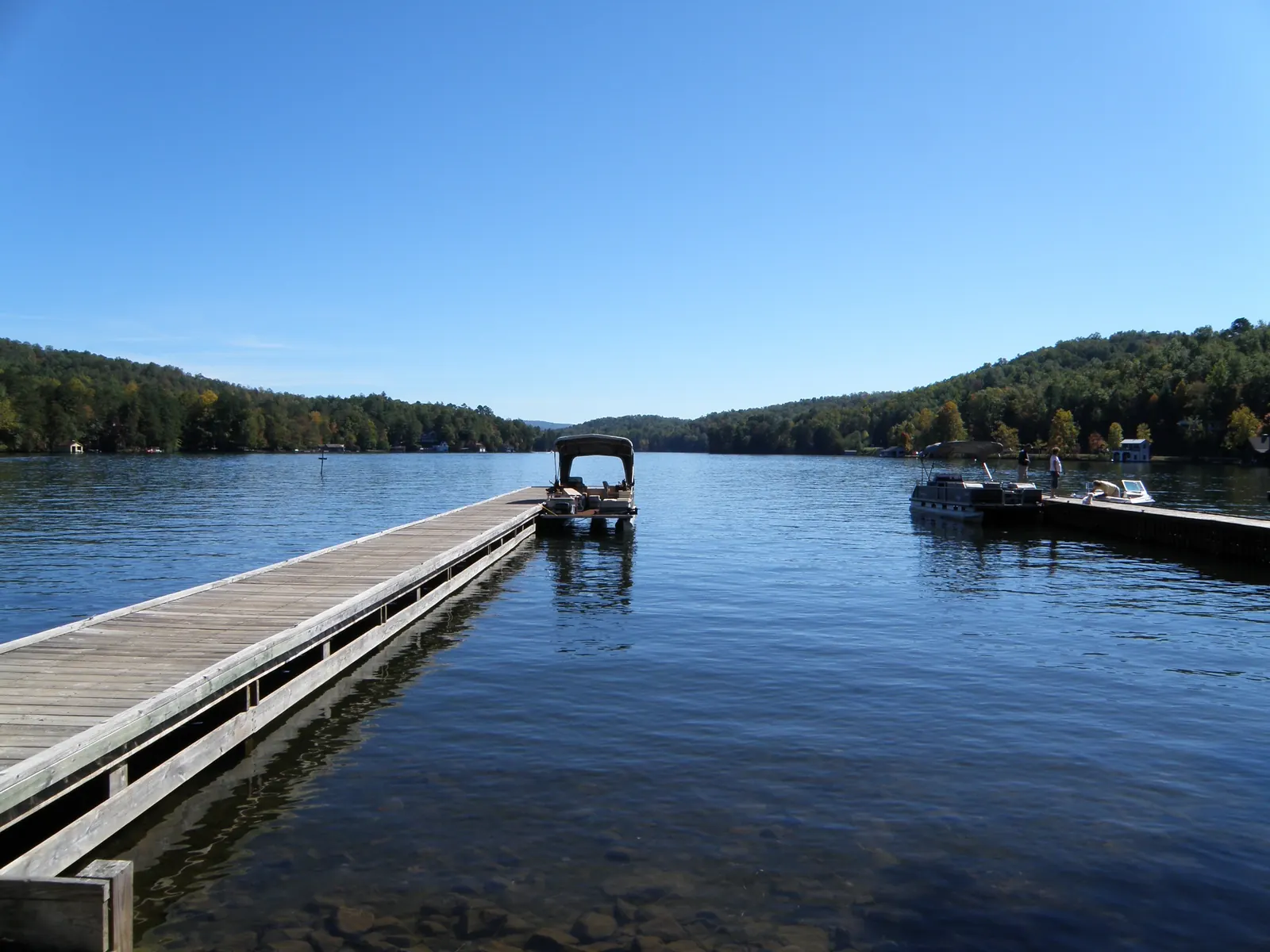 A long wooden dock stretches out over clear, deep-blue lake water from the left, with a pontoon boat at its end and boats moored at a second dock, wooded shores and hills beyond under a cloudless blue sky.