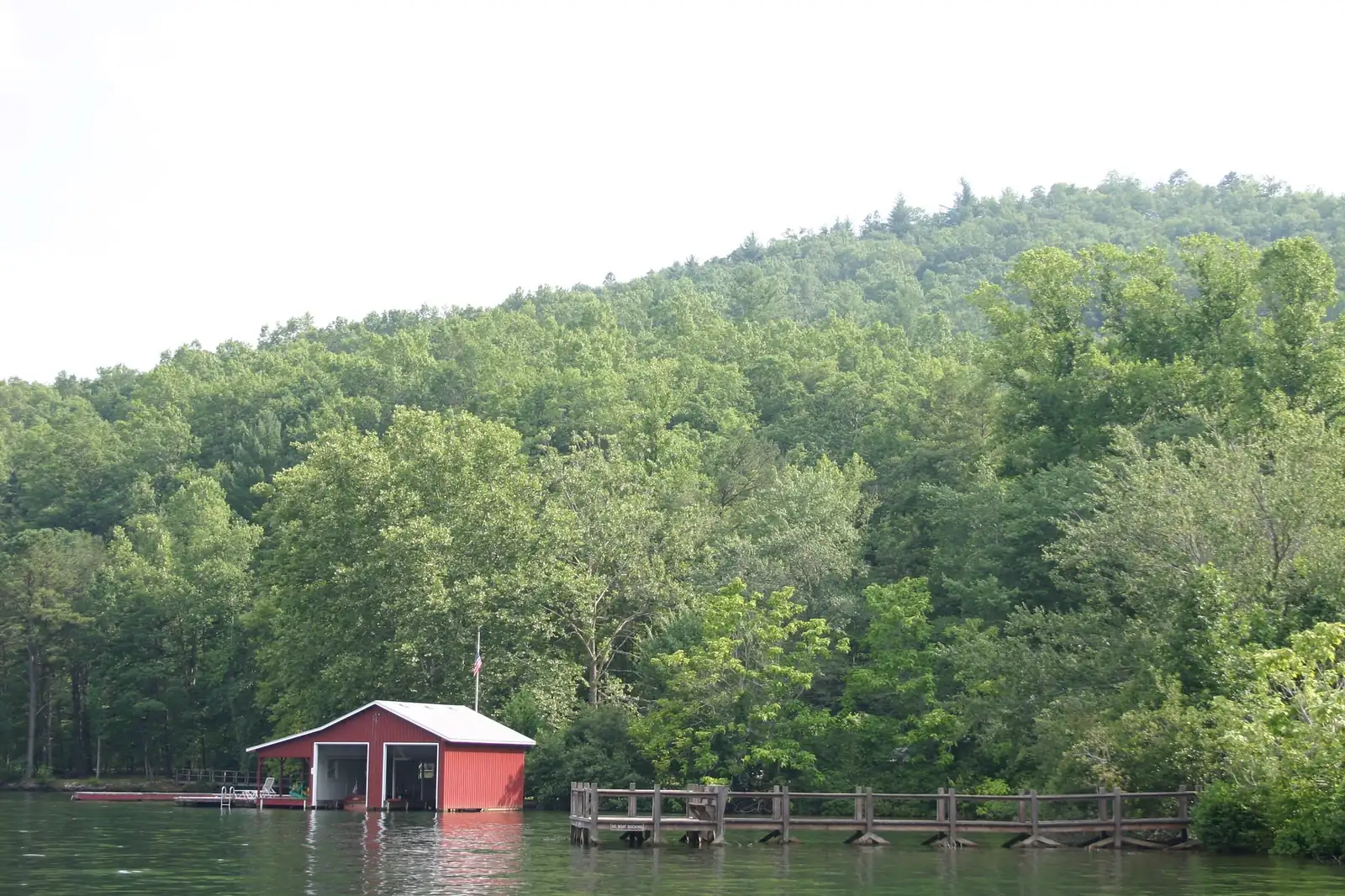 A red boathouse with a white roof and a long wooden dock at the water's edge below a densely wooded green hillside, under a hazy white sky.