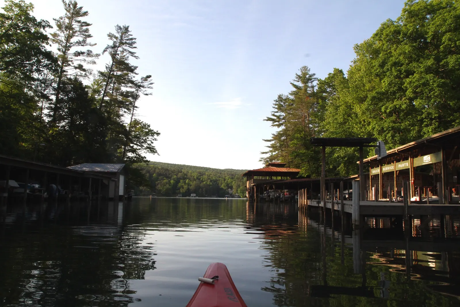 View over the red bow of a kayak gliding down a calm cove between wooden boathouses and covered docks, tall pines and green trees reflected in the dark water.