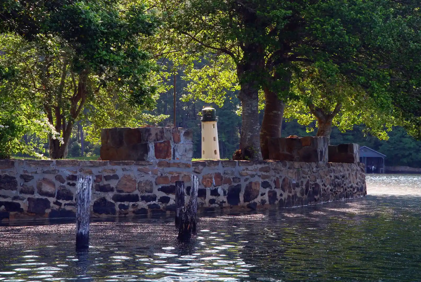 A stone seawall on a shaded point with a small white lighthouse-style tower among old trees, weathered wooden pilings in the water and a boathouse behind.