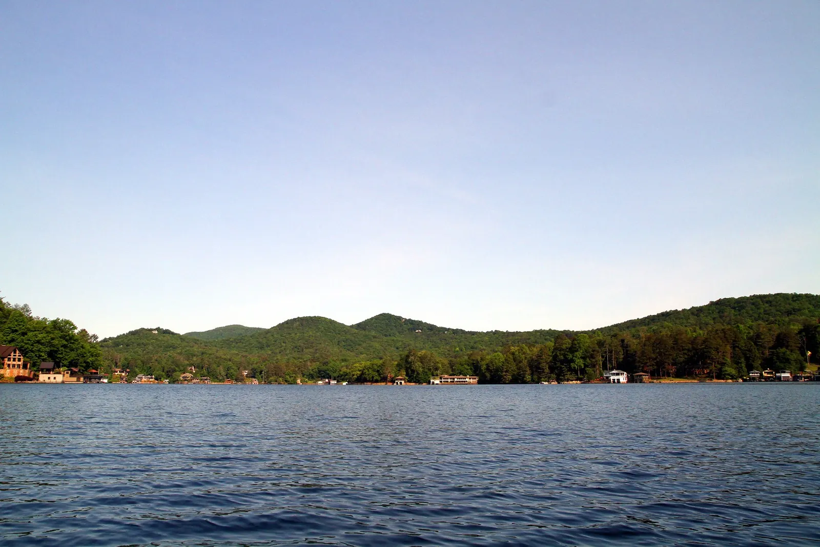Open blue lake water in the foreground stretching to a far shore of wooded rolling hills dotted with lakeside houses and boathouses, under a clear pale-blue sky.
