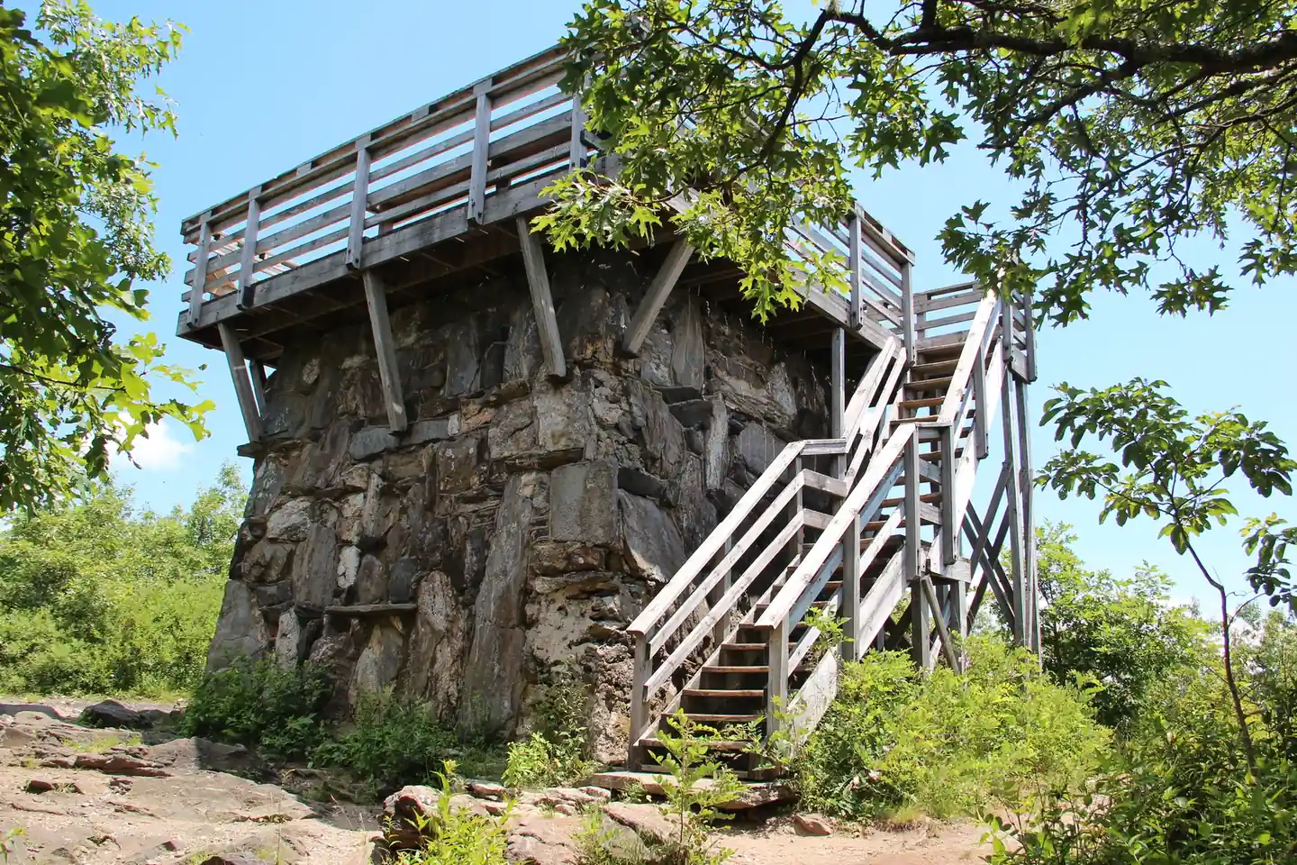 A stone fire-lookout tower base with a wooden observation deck and zig-zag timber staircase, framed by oak leaves under a bright blue sky.