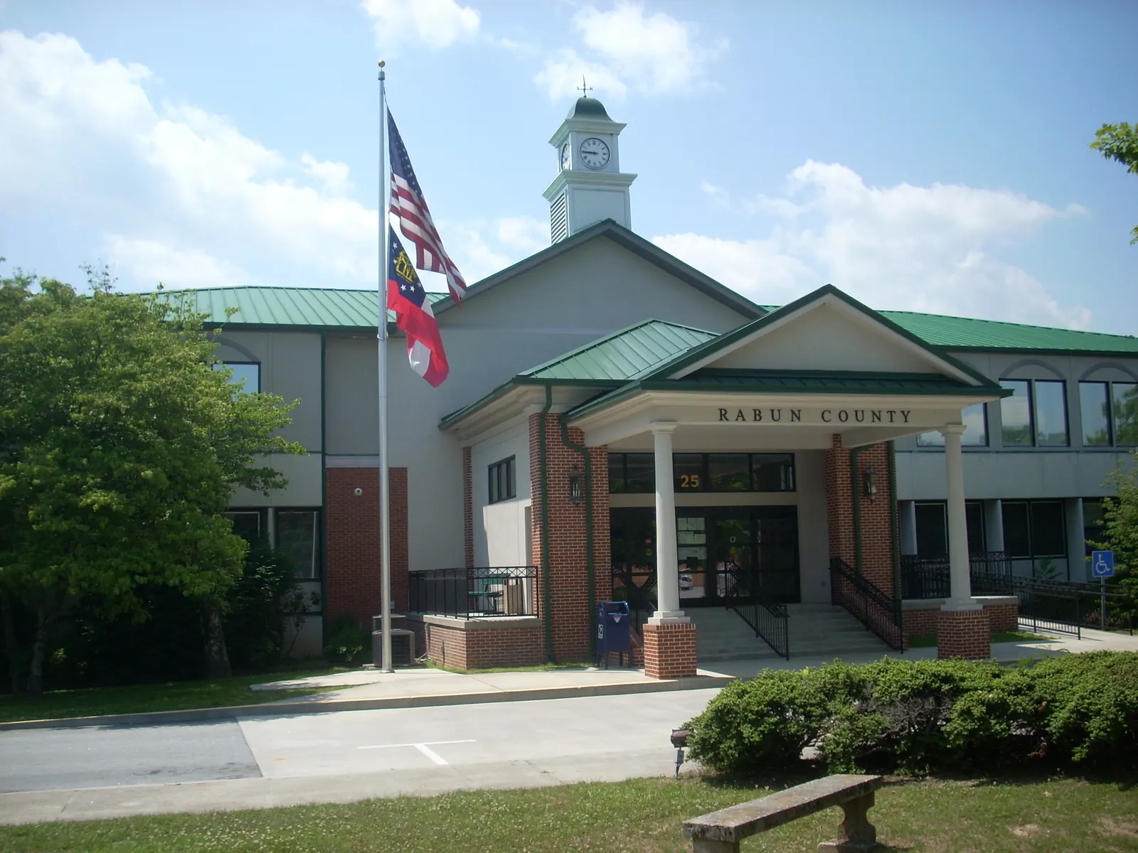 Front of a modern brick-and-stucco county courthouse with a white-columned entry portico lettered "RABUN COUNTY", green metal roofs and a white clock cupola, U.S. and Georgia flags on a pole, a lawn, shrubs and a bench in the foreground under a blue sky with clouds.
