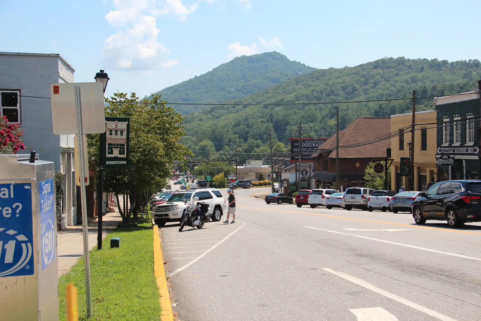 Sunny view down a downtown street: a green "WELCOME / DOWNTOWN" lamp-post banner, a sidewalk under a crape myrtle, angle-parked SUVs and a motorcycle, two-story storefronts and an "Allison Richardson Insurance" billboard on the right, and a forested mountain rising behind the town; a blue "Signal Failure? Call 511" utility box fills the lower-left corner.