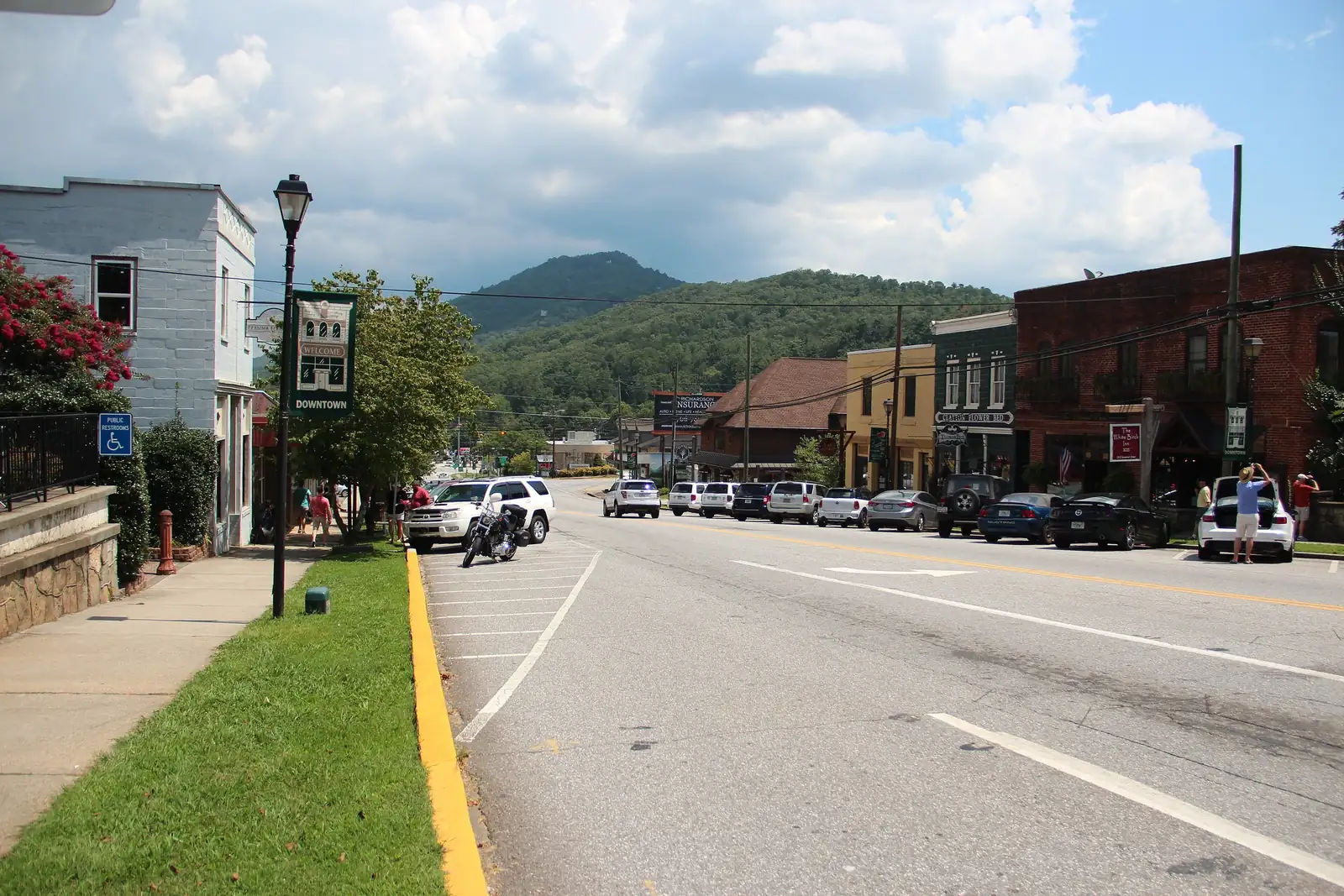 Wide street view of a line of parked cars in front of downtown storefronts: a yellow stucco building, the dark-green "Clayton Flower Bed" building and a red-brick building with a "The White Birch Inn" hanging sign and a "Downtown" banner, under a wooded mountain and a partly cloudy sky; a few visitors with cameras stand by a car at right.