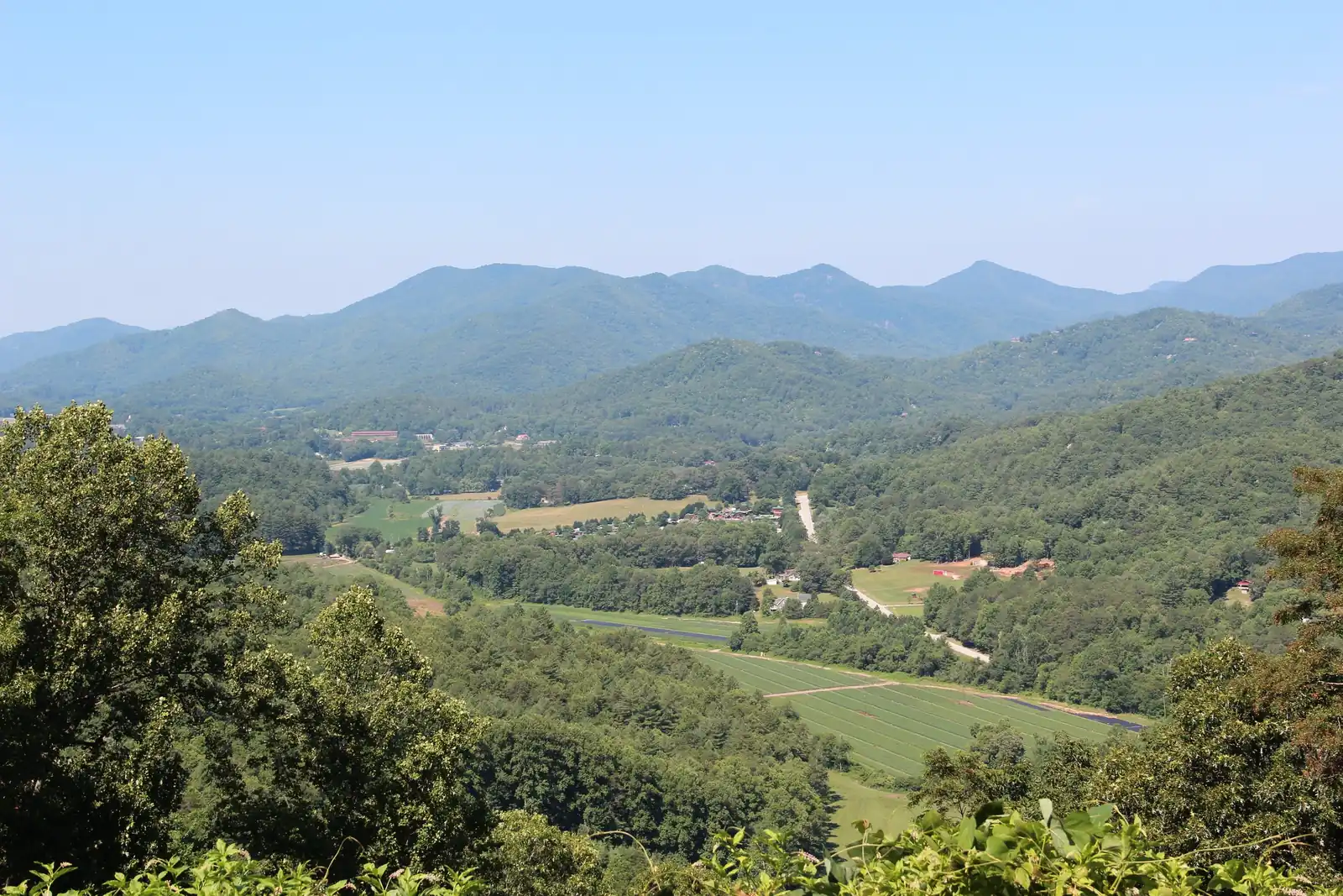 A wide green valley of fields and scattered farms below forested hills, with a long blue mountain ridge across the horizon under a hazy sky.