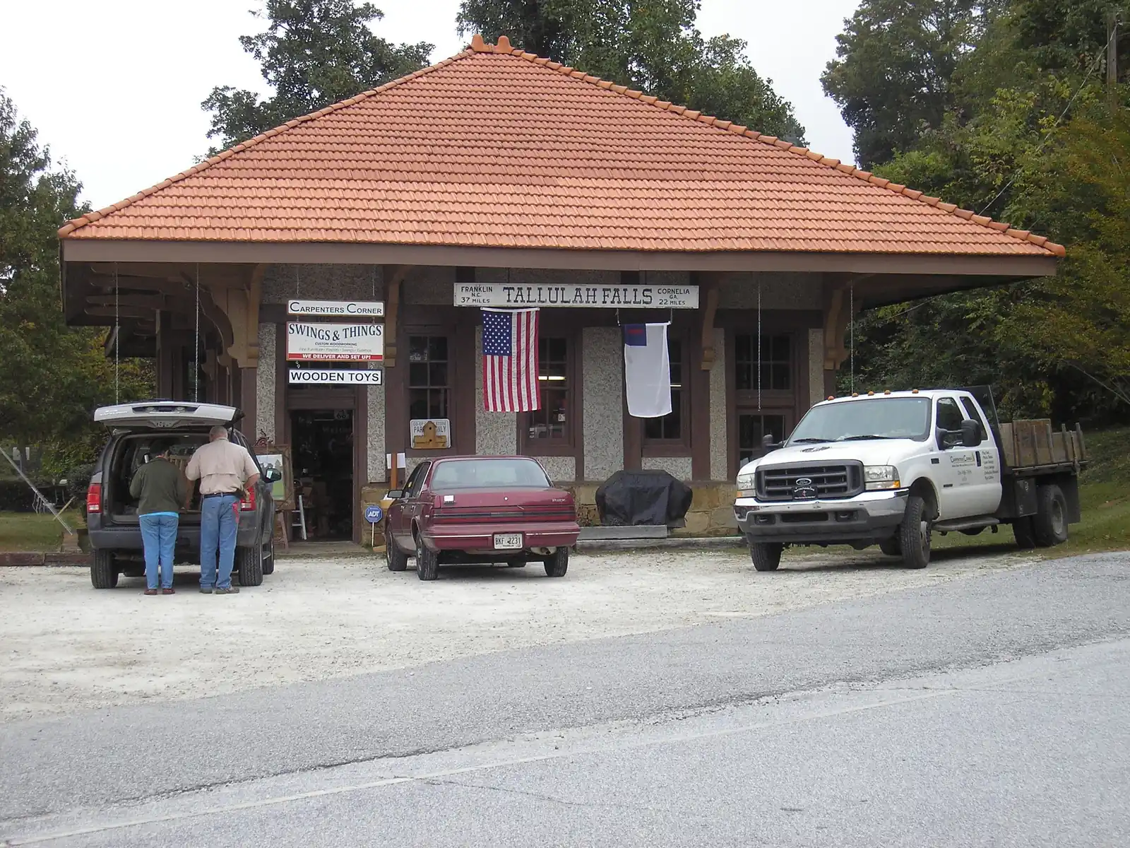 A one-story stucco railroad depot with a red tile hip roof and a sign reading "FRANKLIN N.C. 37 MILES - TALLULAH FALLS - CORNELIA GA. 22 MILES", shop signs for "Carpenters Corner", "Swings & Things" and "Wooden Toys", flags hanging from the eaves, and parked cars and pickups in front.