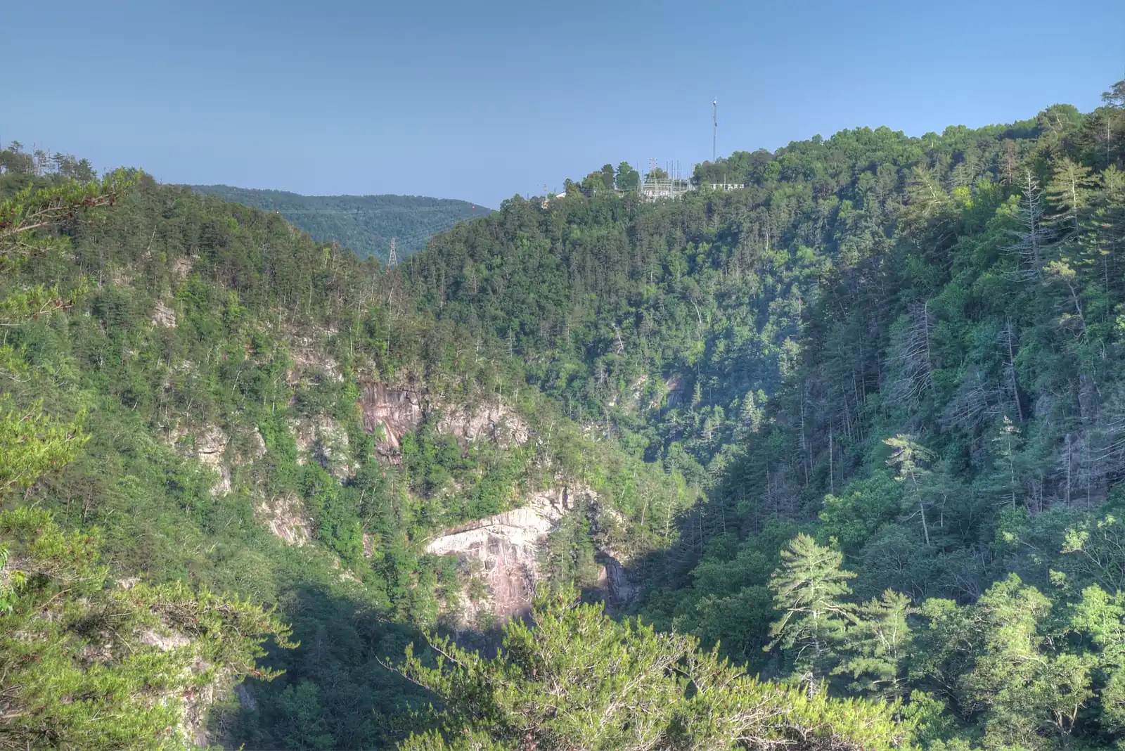 Sunny view across a gorge: steep forested walls with exposed pink-gray granite cliffs, a pale cascade glimpsed low at right, a radio tower on the far rim and a clear blue sky.