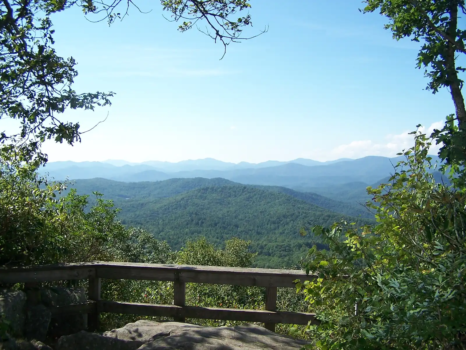 A rustic wooden railing on a granite ledge frames a view through leafy branches across layered blue-green mountain ridges under a clear blue sky.